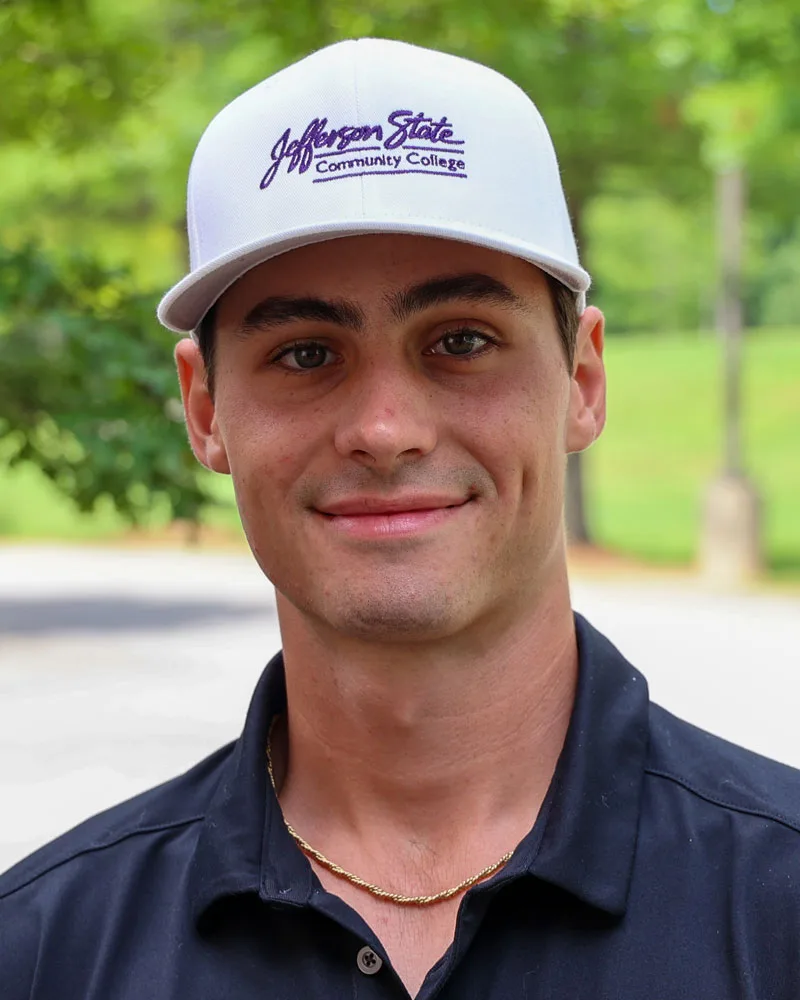 A young man wearing a white Jefferson State Community College cap, a black collared shirt, and a gold chain smiles outdoors with green trees and a blurred background.
