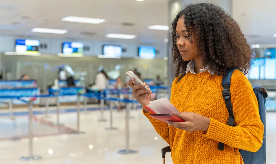 A young woman with curly hair, wearing an orange sweater and a backpack, looks at her phone while holding a passport and boarding pass in an airport check-in area with people and counters in the background.