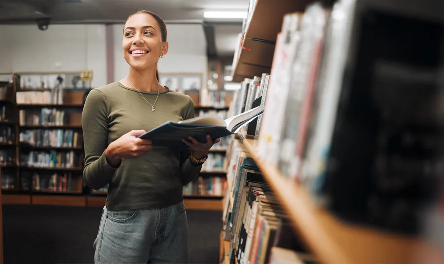 A smiling woman holding an open book stands between bookshelves in a library, looking upward as she browses the collection.