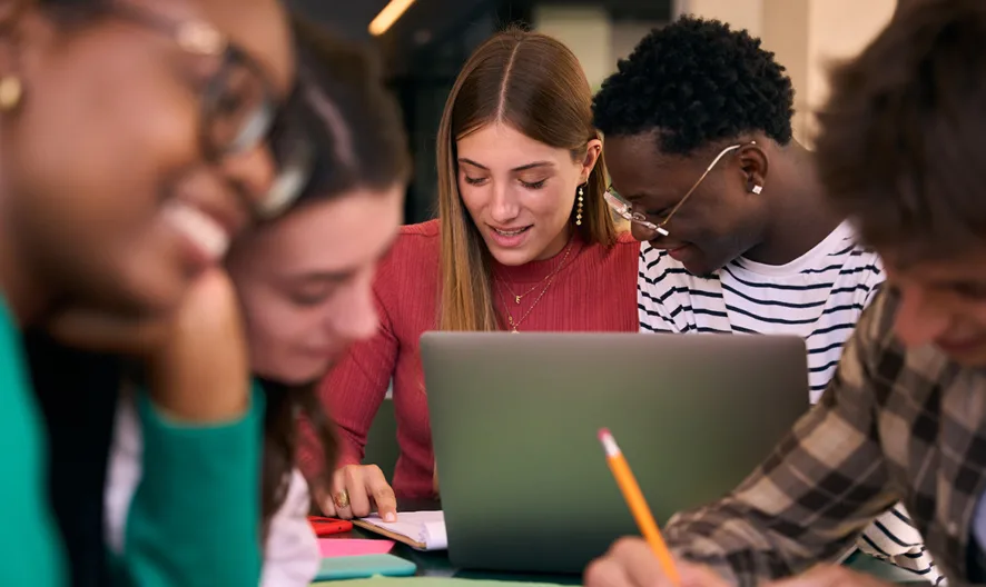 A group of five students sit together at a table, studying and working on assignments. They are focused, with one using a laptop, and others writing in notebooks, suggesting a collaborative and engaged learning environment.