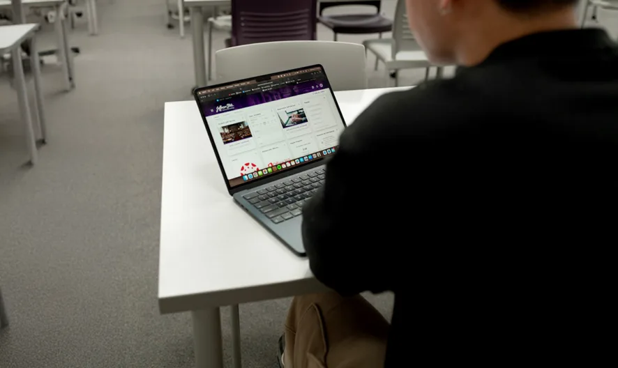 A person sits alone at a desk in a classroom, using a laptop. The classroom has several empty white desks and chairs arranged in rows. The laptop screen displays a website.