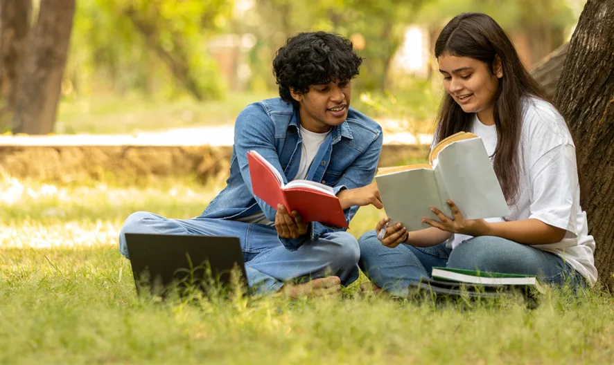 Two young adults sit on grass under a tree, studying together with books and a laptop in front of them, smiling and enjoying an outdoor learning session in a park.