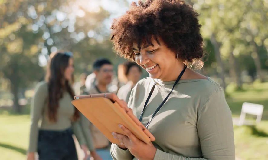 A smiling woman with curly hair uses a tablet outdoors in a sunny park. Three people stand in the blurred background, and trees and grass surround them.