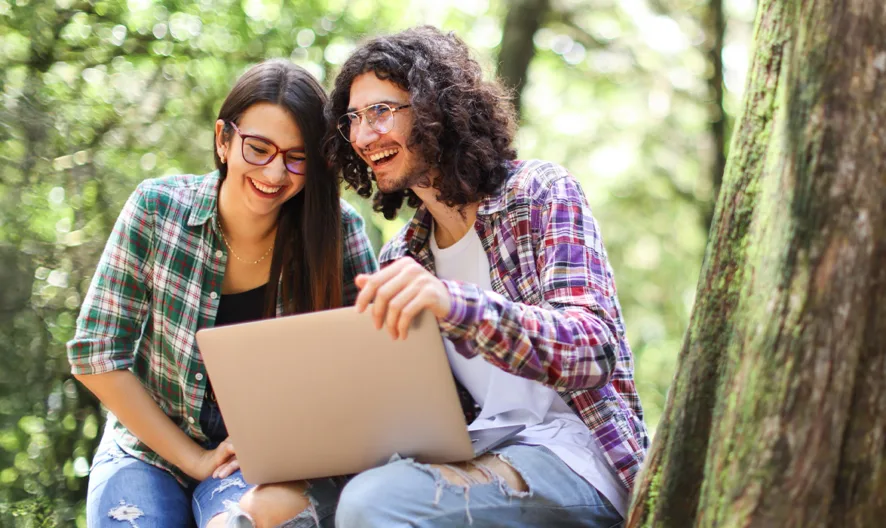 Two young adults sit outdoors by a tree, smiling and looking at a laptop together. They both wear glasses and casual clothes, and the background is filled with green, sunlit trees.