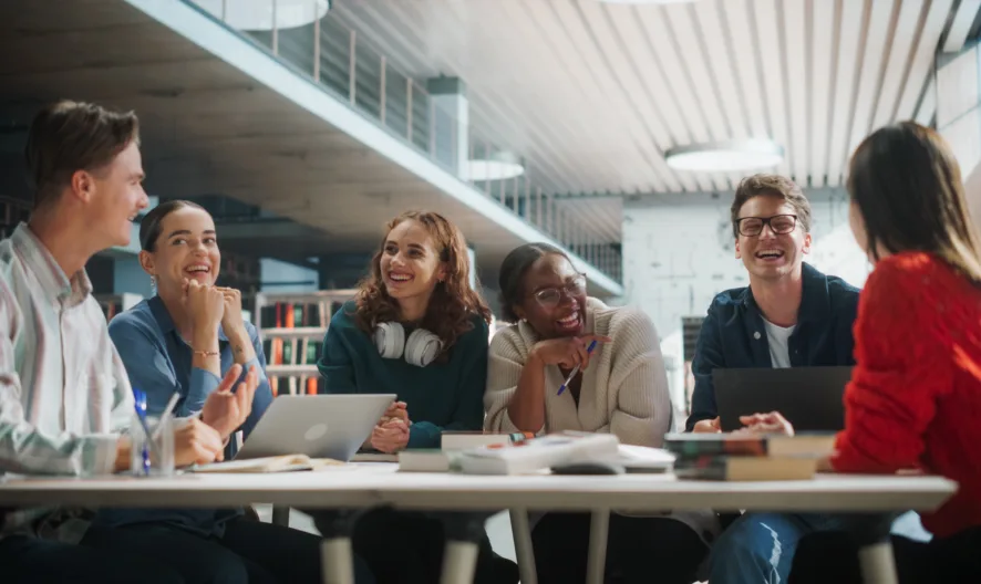A group of six young adults sit around a table in a modern library, laughing and talking. Laptops, notebooks, and books are on the table, and bookshelves are visible in the background.