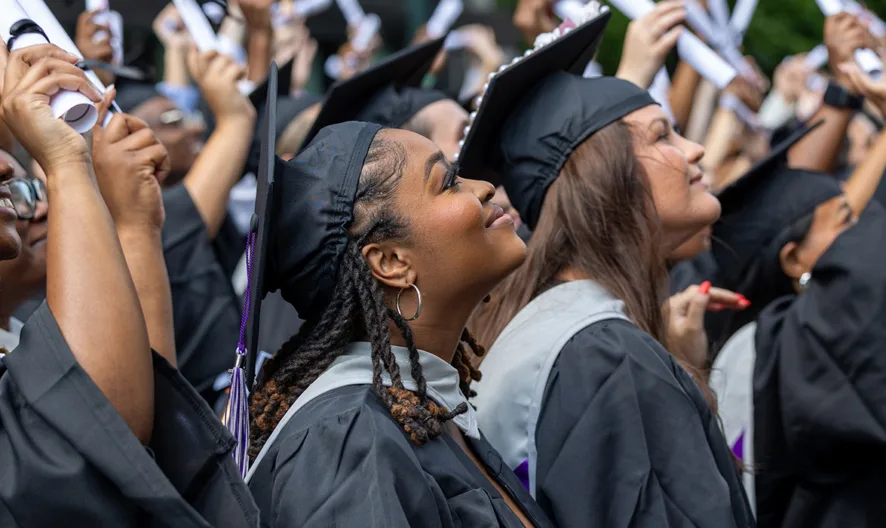 A group of graduates in caps and gowns look upward and celebrate, holding their diplomas and smiling during a commencement ceremony.