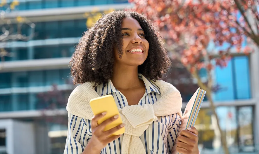 A young woman with curly hair smiles while holding a yellow phone and a notebook outdoors. She wears a striped shirt with a sweater draped over her shoulders, with modern buildings and trees in the background.