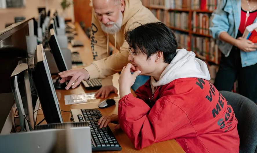 A student in a red jacket uses a desktop computer in a library while an older man assists. In the background, two other people stand by the bookshelves, one holding books and the other reading.