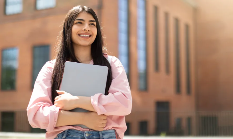 A young woman with long dark hair stands outside a modern brick building, smiling and holding a closed laptop in her arms. She is wearing a pink shirt and jeans, and the sun is shining brightly.