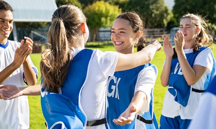 A group of teenagers in sports uniforms smile and celebrate on a grassy field, with two girls hugging while teammates clap and cheer in the background.