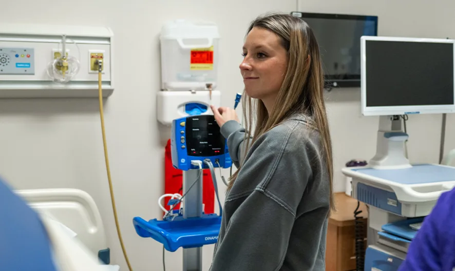 Two women are in a hospital room, one adjusting a medical monitor while the other looks down. There is a hospital bed, medical equipment, and a computer in the background.