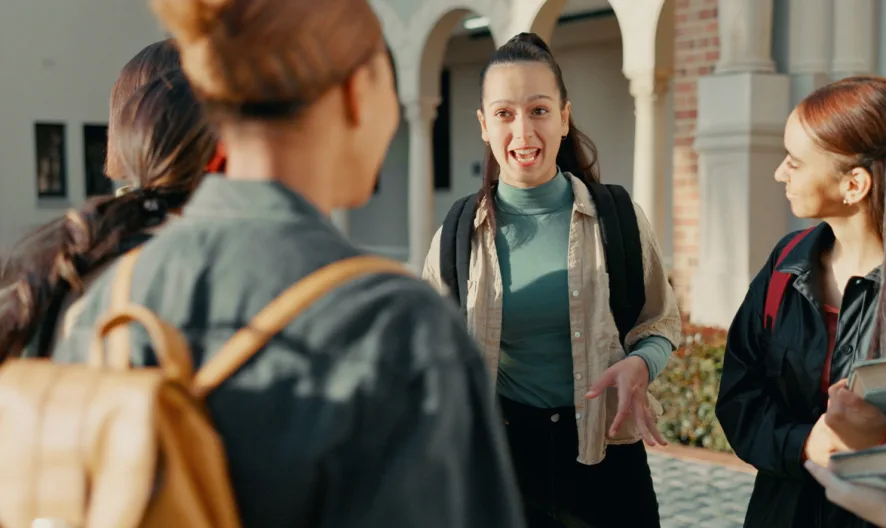 A group of five young adults stand outside a building, talking and smiling. One person in the center gestures while others listen, holding books and backpacks, suggesting a friendly conversation among students.