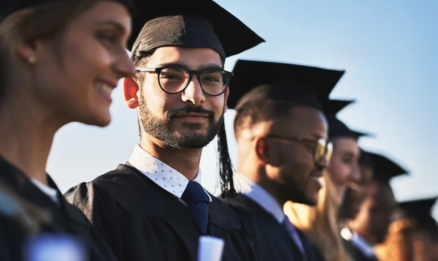 A group of graduates in caps and gowns stand in a row outdoors, smiling and looking ahead, with one person in the center wearing glasses and facing the camera. The sky is clear and sunny.