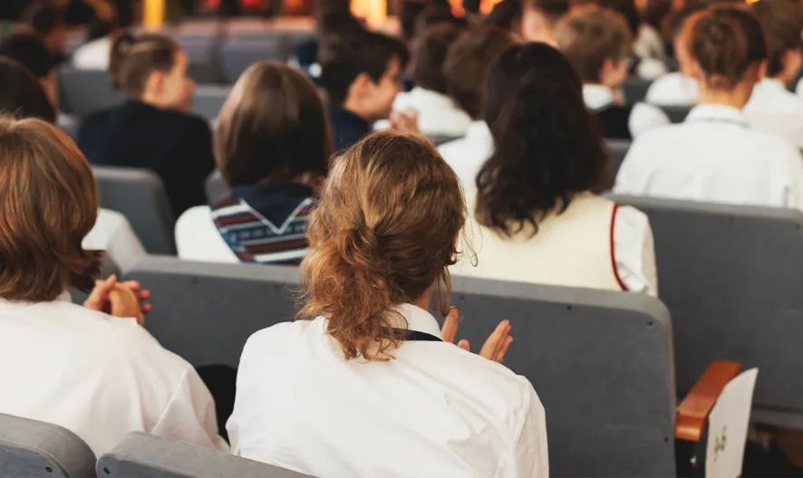 A group of students in school uniforms sit in rows of gray chairs in an auditorium, facing forward and listening attentively to an unseen speaker or presentation.
