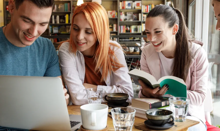 Four young adults sit at a table in a cozy cafe, smiling and gathered around a laptop. Books, notebooks, and drinks are on the table, suggesting they are studying or collaborating on a project together.