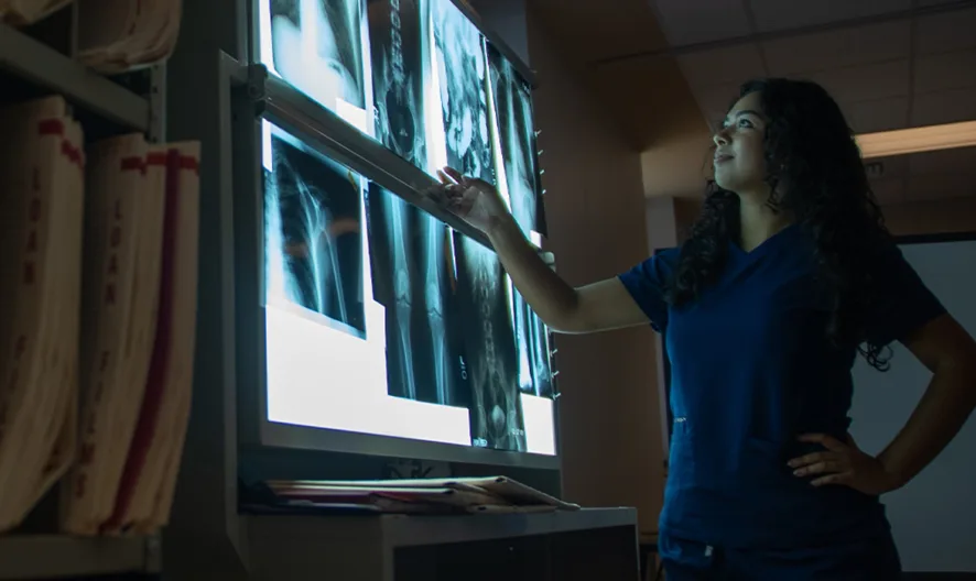 A person in medical scrubs examines X-ray images on a lightbox in a dimly lit room, with shelves of files visible nearby.