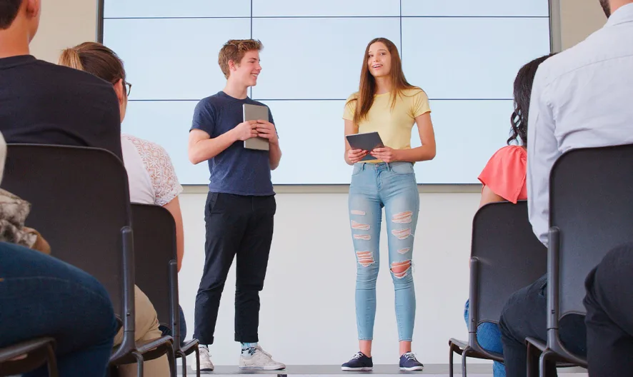 Two young adults stand on a stage holding tablets, presenting to an audience seated in rows, with a large screen behind them. The presenters are smiling and casually dressed.
