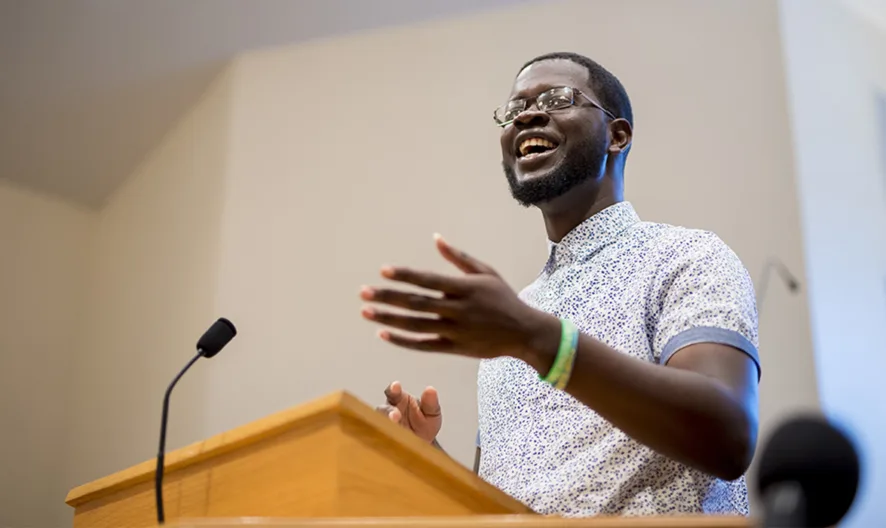A man wearing glasses and a patterned shirt speaks passionately at a podium, gesturing with one hand, in a well-lit indoor setting with a microphone nearby.