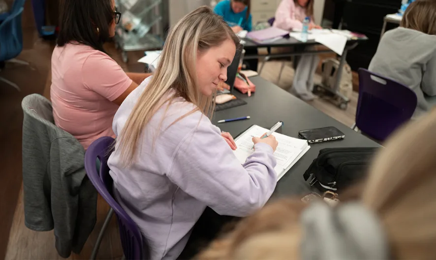 A student sits at a desk in a classroom, writing in a notebook, surrounded by other students working at computers and tables.