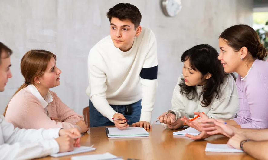 Six young adults sit around a table with notebooks, actively discussing and collaborating. One person stands, leaning in and speaking, while the others listen and engage in conversation.