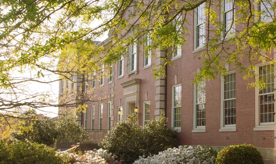 A brick building with tall windows is partially shaded by leafy green trees. Sunlight shines through the branches, illuminating bushes and flowering plants along the walkway.