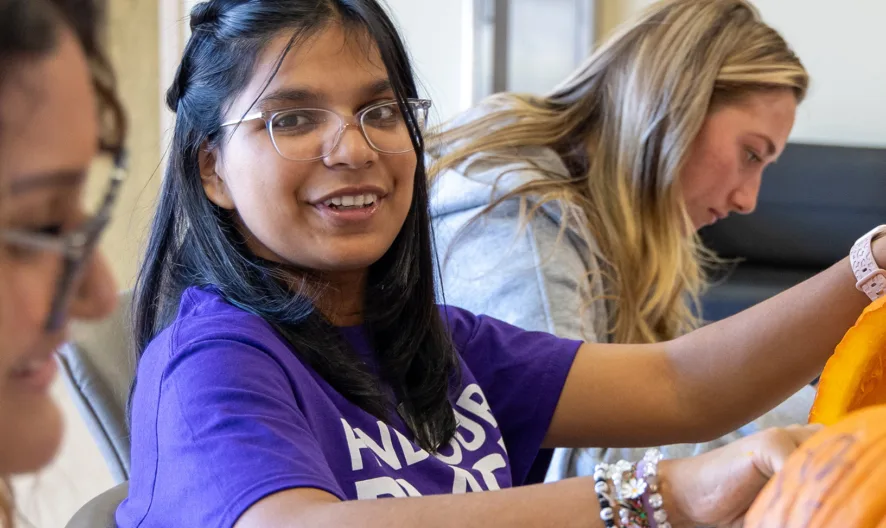 Three people sit at a table carving pumpkins. The person in the center, wearing glasses and a purple shirt, smiles at the camera while working on a pumpkin. The others are focused on their pumpkins.