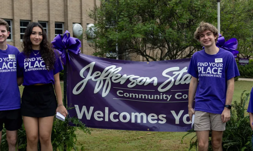 Four young people in matching purple Find Your Place at Jefferson State shirts stand smiling in front of a “Jefferson State Community College Welcomes You” banner outdoors. A booth and balloons are visible in the background.