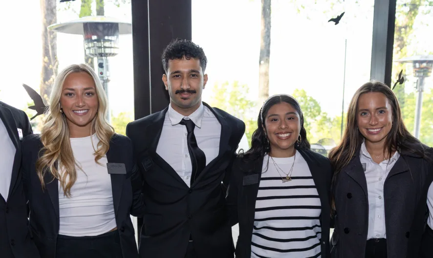 Six young adults dressed in professional attire stand side by side indoors, smiling at the camera. Natural light filters in through large windows behind them, showing green trees outside.