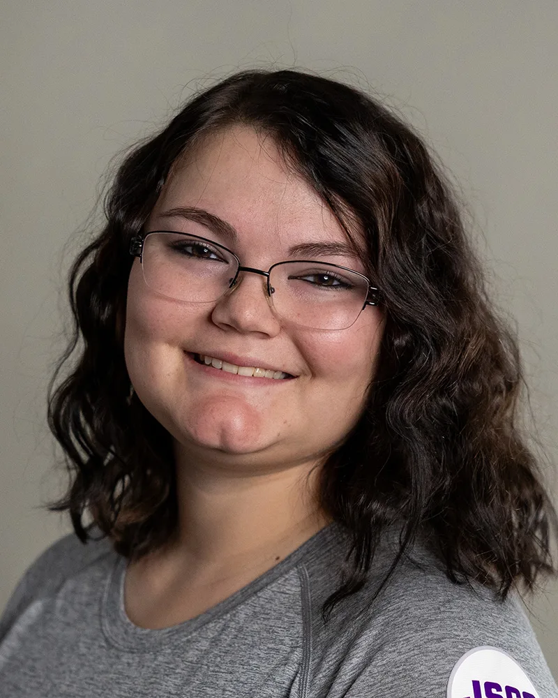 A person with wavy brown hair wearing glasses and a gray top smiles at the camera against a plain background.