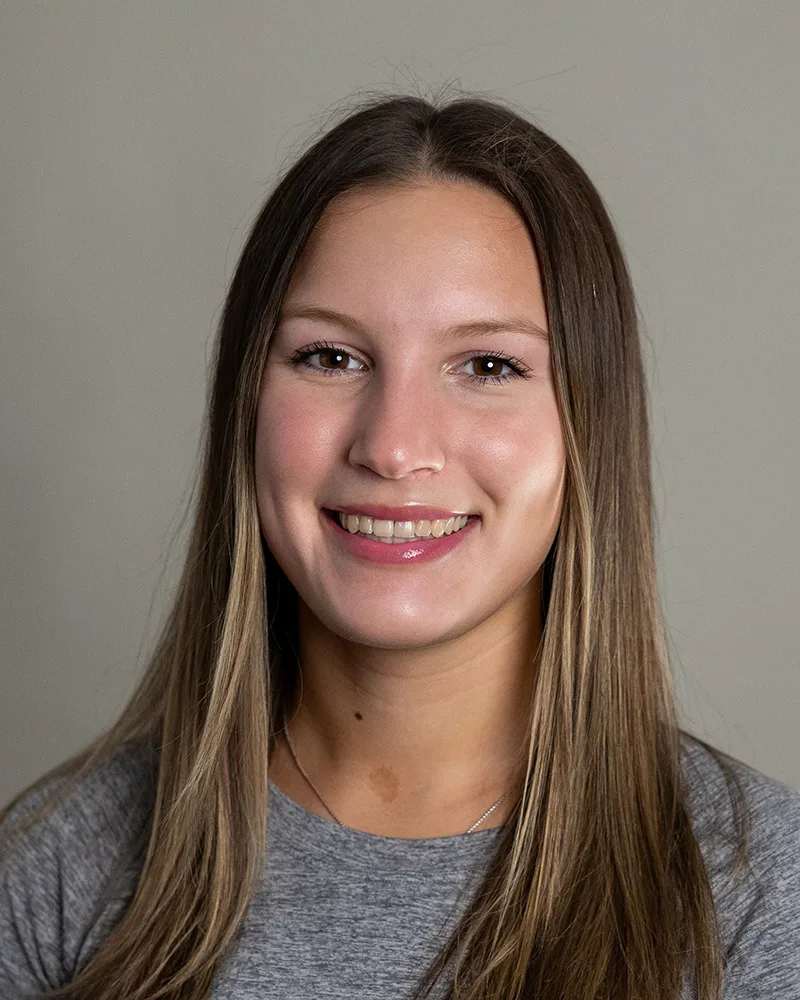 A young woman with straight brown hair, wearing a gray top, smiles at the camera against a plain light background.