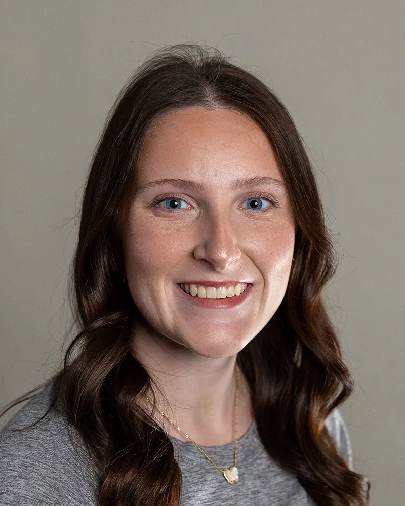A woman with long brown wavy hair and blue eyes smiles at the camera. She is wearing a grey top and a gold necklace with a pendant, posed against a plain light background.