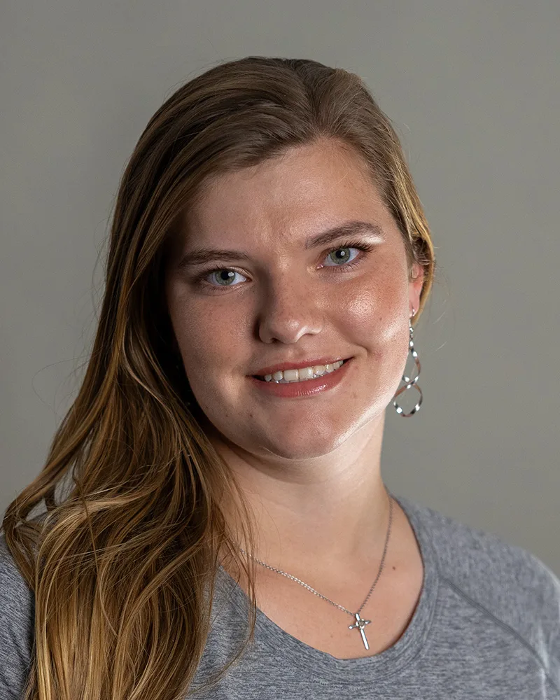 A young woman with long, light brown hair smiles softly. She is wearing a gray top, a silver necklace with a cross pendant, and large hoop earrings, with a plain gray background behind her.