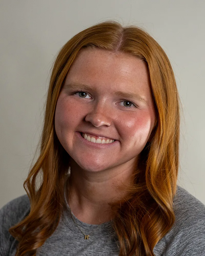 A young woman with long red hair and fair skin smiles at the camera. She is wearing a gray top and a necklace with a small pendant. The background is plain and light-colored.