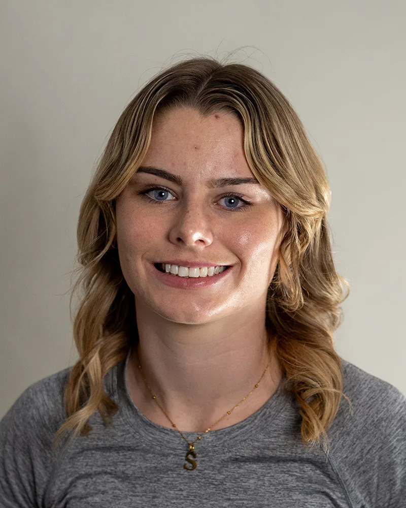 A young woman with wavy blonde hair wearing a gray shirt and a gold necklace with an S pendant smiles at the camera against a plain light background.