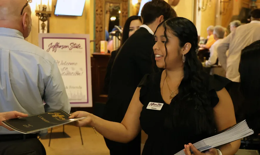 A woman in a black dress hands out programs to guests at an indoor event. People are lined up, some holding programs, in a warmly lit ornate venue. Everyone appears engaged and happy.