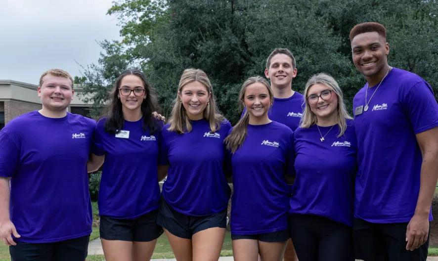 Seven smiling young adults wearing matching purple shirts stand outdoors in a row with their arms around each other, in front of a brick building and trees on a cloudy day.