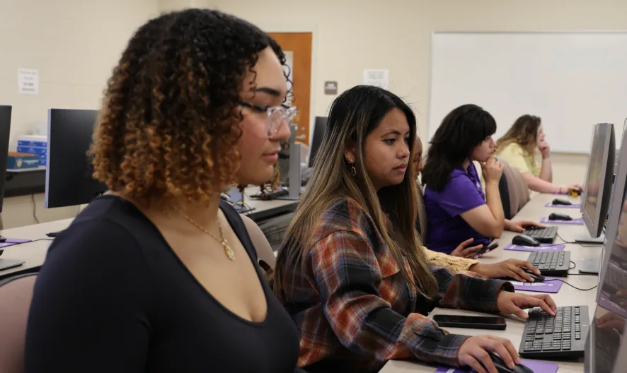 Four students sit in a row at computer desks in a classroom, focused on their desktop monitors and using keyboards and mice. The classroom has beige walls and fluorescent lighting.