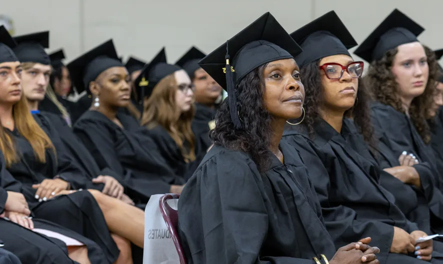 A group of graduates in black caps and gowns sit in rows at a ceremony, facing forward and listening attentively. Some have tassels on their caps and are holding programs.