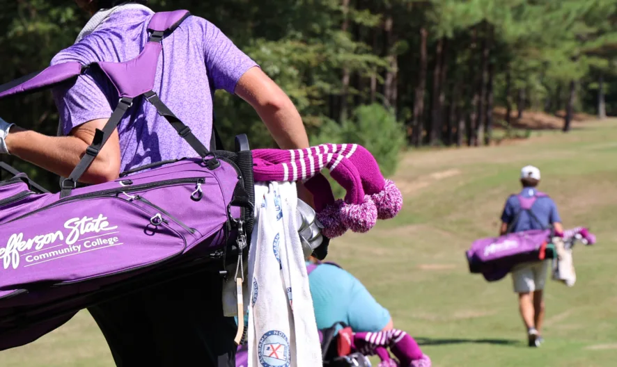 Golfers carrying purple Jackson State Community College golf bags walk across a grassy course bordered by trees on a sunny day. The focus is on one golfer walking in the foreground.