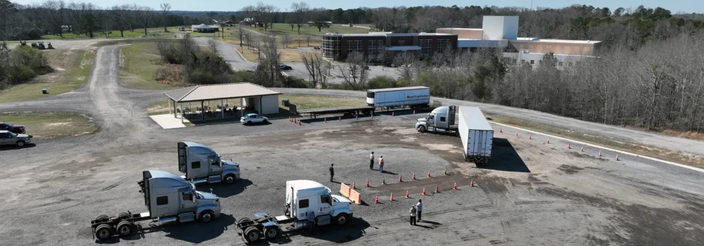 Aerial view of a truck driving practice area with three trucks, orange cones arranged for maneuvering, a few people supervising, a covered shelter, and a large building surrounded by trees in the background.