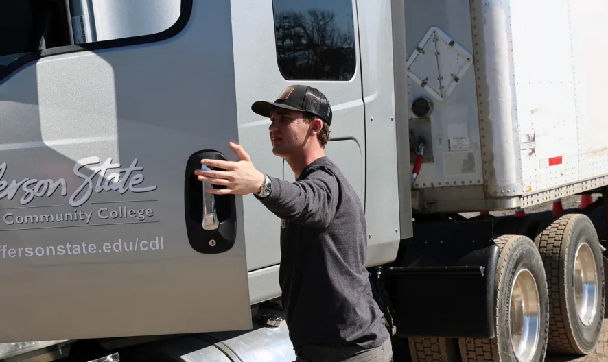 A man wearing a cap opens the door of a large truck labeled Jefferson State Community College near several orange traffic cones.