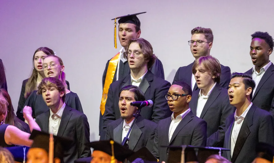 A diverse group of choir singers, some in graduation attire, perform on stage as a conductor leads them. The singers wear black suits or gowns and stand in front of microphones with sheet music visible below.