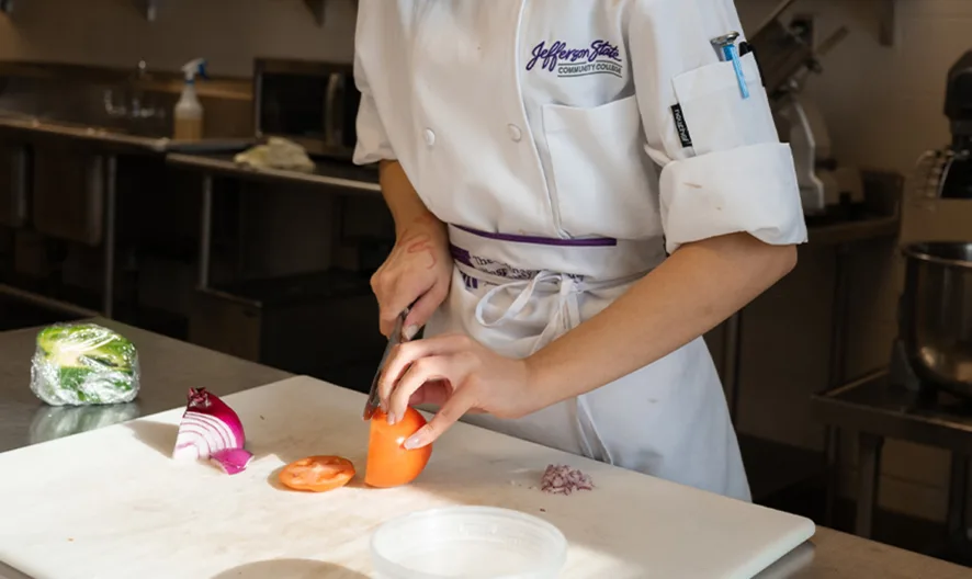 A person in a white chefs uniform slices a tomato on a cutting board in a professional kitchen, with chopped onions, containers, and utensils nearby.
