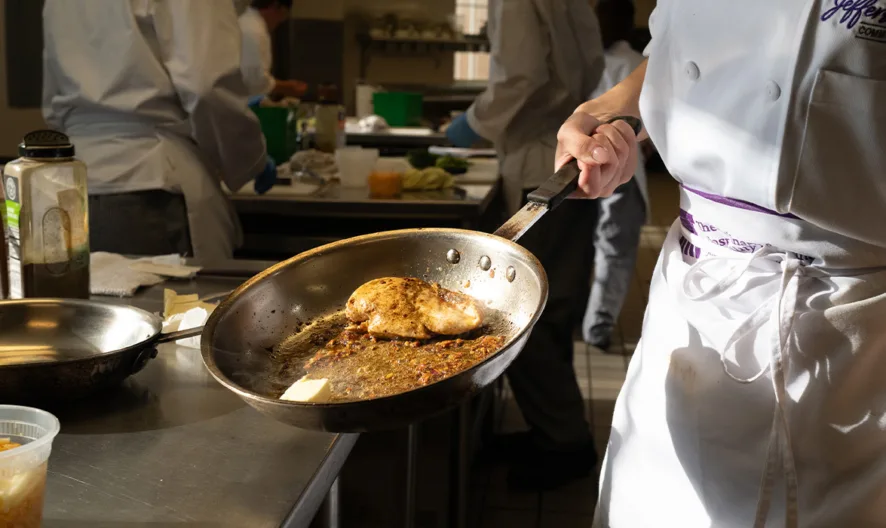 A chef in a white uniform and apron cooks a piece of chicken breast in a skillet on a stainless steel countertop in a bright, busy kitchen. Other chefs and cooking ingredients are visible in the background.