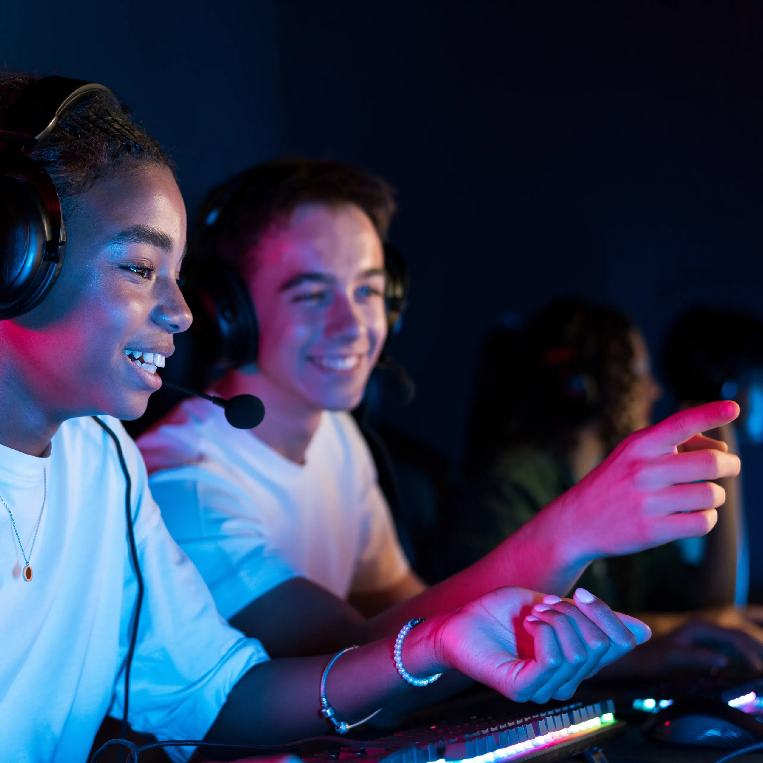 Two young people wearing headsets sit at computers, smiling and focused, as one points at the screen. They are illuminated by colorful lighting, suggesting they are playing a video game together in a gaming setup.