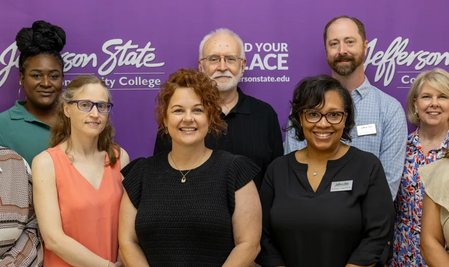 A group of twelve people smiling and standing together in front of a purple Jefferson State Community College banner. The group is diverse in age, gender, and ethnicity, and many wear name badges.