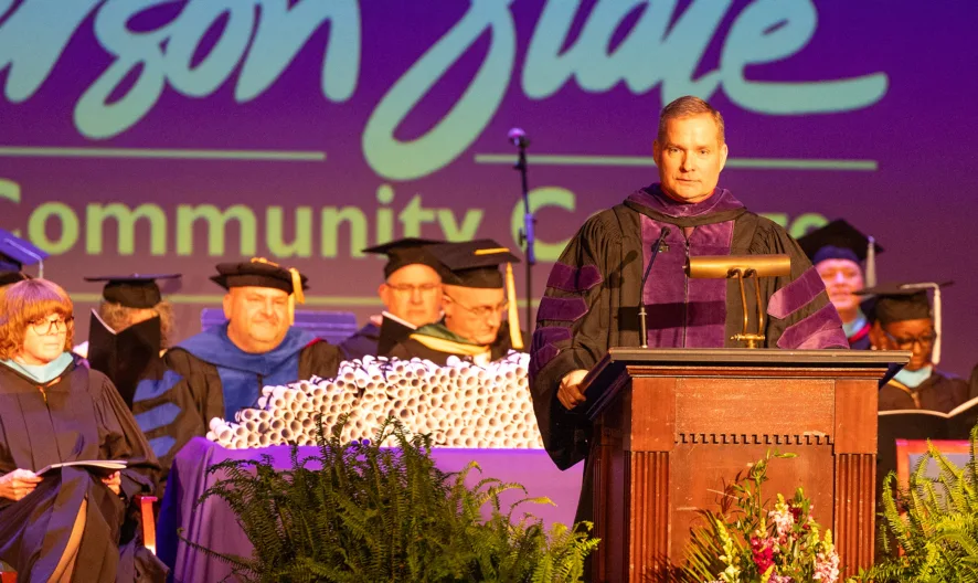 A person in academic regalia speaks at a podium during a graduation ceremony, with faculty seated behind and a “Jefferson State Community College” sign displayed on a purple-lit stage.