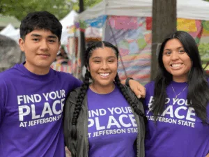 Three smiling young adults wearing matching purple Find Your Place at Jefferson State T-shirts stand together outdoors, posing with arms around each other in front of colorful tents.