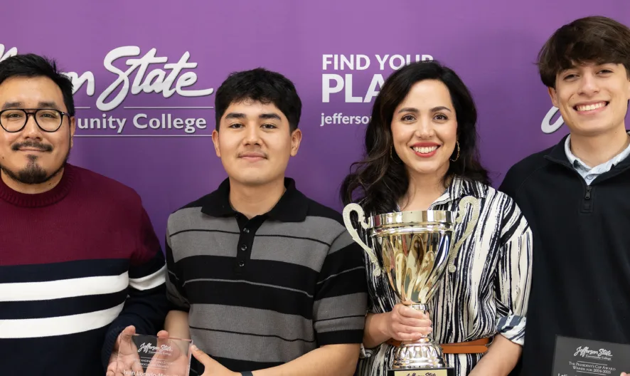 Four people stand in front of a purple Jefferson State Community College backdrop. Two hold glass awards, and one holds a large trophy. All are smiling and dressed in casual or business-casual attire.