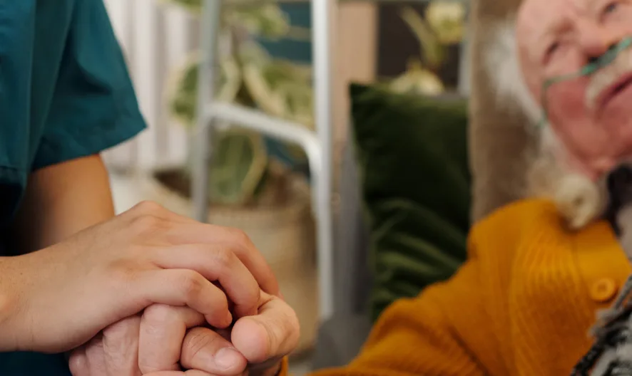 A healthcare worker holds the hand of an elderly patient lying in bed with an oxygen tube, offering comfort and support.
